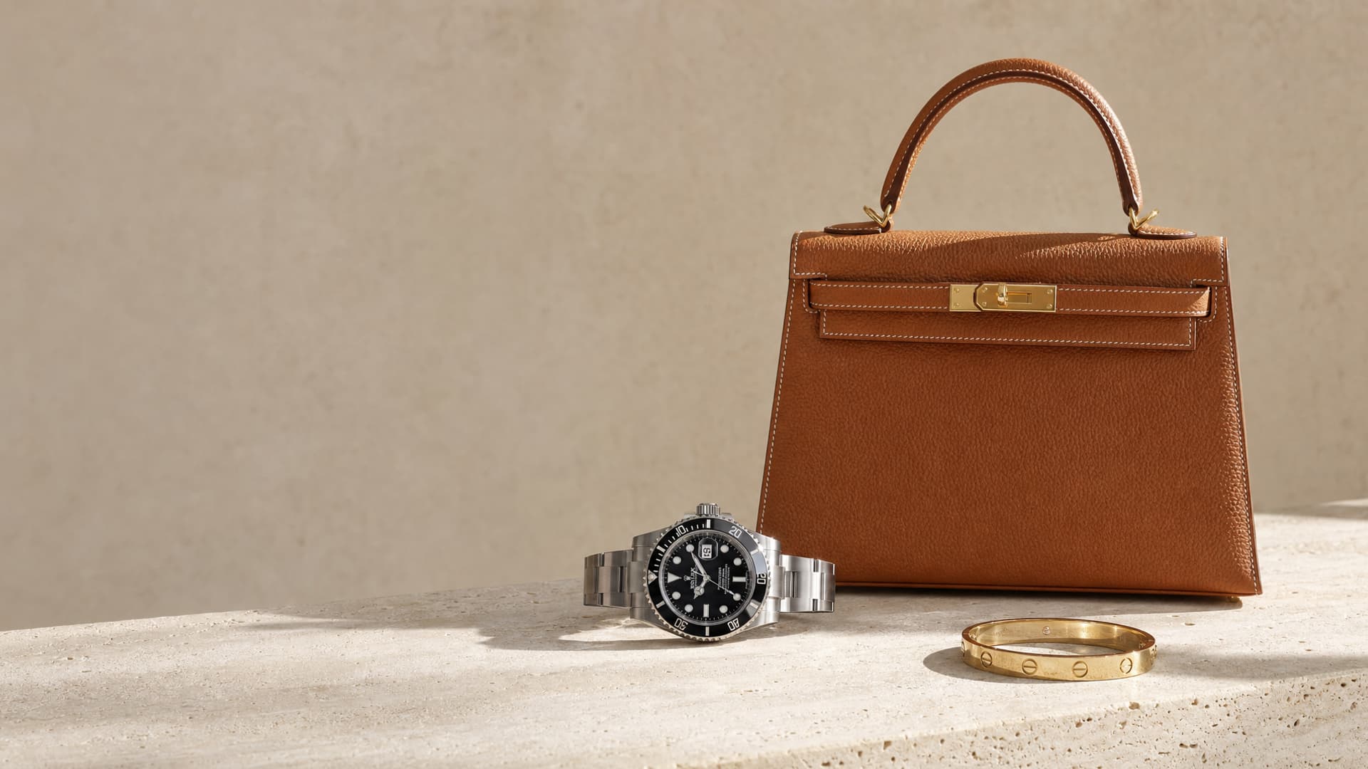 Sac Kelly Hermès, montre Rolex Submariner et jonc Cartier Love posés sur du travertin